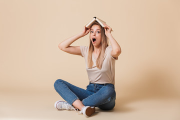 Poster - Photo of amusing funny woman wearing casual clothing sitting on floor with legs crossed and putting open book on head, isolated over beige background