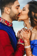 Wind blows around Hindu groom in blue sherwani and bride in lehenga posing in white house with gorgeous seaside view behind them