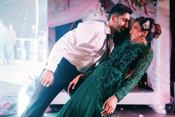 Canvas Print - Indian groom in a classy black suit and beautiful bride in a green evening gown dance in the restaurant