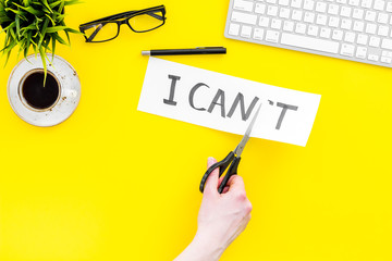 Self confidence concept. Hands cutting the letter t of written word I can't by sciccors. Office desk. Yellow background top view copy space