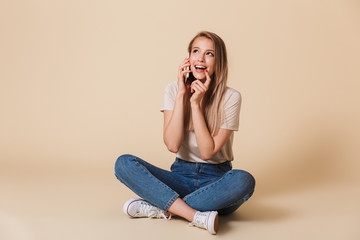 Sticker - Image of pleased sociable woman wearing casual jeans talking on mobile phone while sitting on floor with legs crossed, isolated over beige background