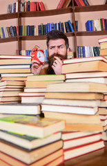 Leinwandbilder - Teacher or student with beard studying in library. Man, scientist peeking out of piles of books with alarm clock. Man on thoughtful face looking at clock, bookshelves on background. Deadline concept.