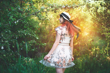 Wall Mural - A beautiful girl with long red hair in a hat walks in a magical garden. Summer vacation concept