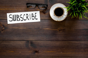 Poster - Email subscribe concept. Hand lettering subcribe on work desk with plant, glasses, cup of coffee on dark wooden background top view copy space