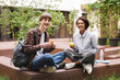 Couple of smiling students sitting on bench with sandwich and green apple and happily looking in camera while spending time together in courtyard of university
