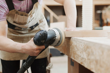 Carpenter polishes the wooden surface of furniture products. Electric eccentric orbital grinding machine. Mechanized woodworking carpenter hand tool in working carpentry workshop.