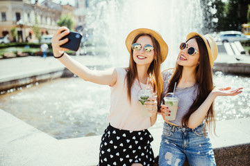 Two cheerful young women pose against fountain with smoothie, make selfie on modern mobile phone or talk online on video call, share their impressions about resort abroad