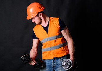 Worker, contractor, builder on busy face with muscular biceps. Builder in helmet working out, training. Man in helmet, hard hat holds heavy dumbbells, black background. Strong builder concept.