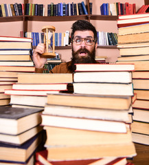 Leinwandbilder - Man on surprised face holds hourglass while studying, bookshelves on background. Time flow concept. Man, scientist in glasses looks at hourglass. Teacher or student with beard studying in library.