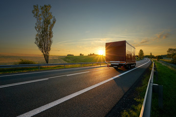 Wall Mural - red small truck driving on asphalt road around farm fields in rural landscape at sunset