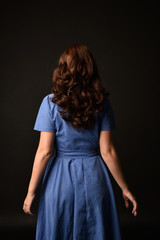 3/4 portrait of brunette lady wearing blue dress, facing away from camera. posed on black studio background.