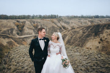 A stylish bridegroom in a black suit and a beautiful bride in a lace dress go and smile on the background of gray rocks and hills. Autumn wedding portrait of cute newlyweds. Film effect.