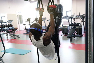 Wall Mural - A black muscular guy in a white T-shirt and headphones hangs on the gym rings and understands his legs.