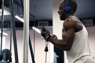 Wall Mural - Emotional photo of a black muscular man in a T-shirt and headphones, shaking hands with a simulator in the gym.