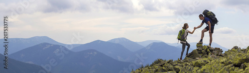 Fotomural Young tourists with backpacks, athletic boy helps slim girl to clime rocky mountain top against bright summer sky and mountain range background