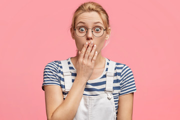 Wall Mural - Terrified female with blonde hair and blue eyes, covers mouth with hand, has worried expression, dressed in casual clothing, stares through spectacles, being puzzled by bad news about health