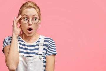 Wall Mural - Indoor shot of stunned blonde woman with blue eyes, keeps mouth wide opened, being puzzled by something, wears denim overalls and sailor t shirt, stands against pink background with blank space