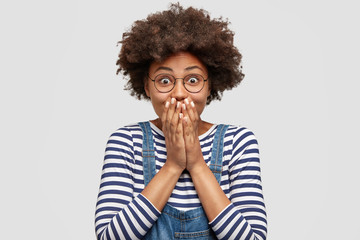 Photo of amazed excited joyful female with Afro hairstyle, covers mouth and exclaims with happiness, expresses excitement and positiveness, dressed in casual clothes, isolated on white wall.