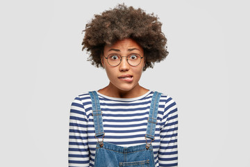 Wall Mural - Embarrassed beautiful African American female bites lower lip nervously, raises eyebrows and has puzzled expression, dressed in casual clothing, isolated over white background. Emotions and ethnicity