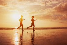 Silhouette Of Two Runners People Jogging On Sunset Sea Beach. Healthy Lifestyle Background With Copyspace. Family Leisure Activities, Sport And Workout. Man And Woman Running.