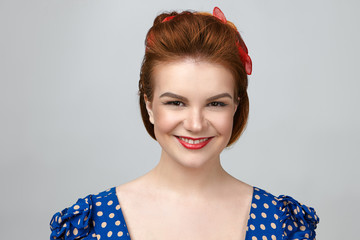 Wall Mural - Happiness, joy and positivity concept. Studio shot of beautiful emotional young red haired Caucasian woman with bright make up and vintage hairdo relaxing indoors, smiling joyfully at camera