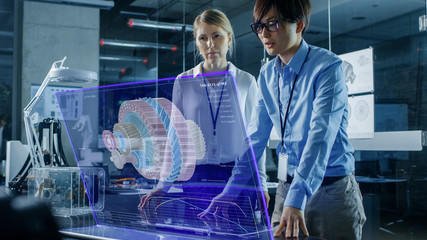 Wall Mural - Male and Female Engineers Have Discussion while Using Modern Computer With Transparent Holographic Display. Monitor Shows Visualization of a Mechanical Gear Part. Shot in Modern Office.