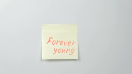 closeup woman's hands stick yellow sticker paper sheets with words forever young.