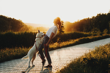 Wall Mural - Full length portrait of beautiful young caucasian woman having fun in the park against sunset while her dog is embracing and playing with her.