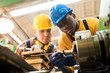 © Seventyfour - Two concentrated workers wearing protective helmets and rubber gloves using lathe in order to machine workpiece, interior of plant production department on background