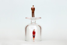 A Miniature Woman In A Glass Cup And A Miniature Man On Top Of A Glass Cup. The Concept Of The Gender Promotion Gap.