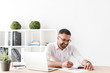 © Drobot Dean - Photo of pleased businessman 30s in white shirt sitting at table and writing down information at notebook, while working with laptop in office