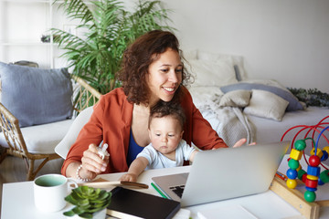 Attractive young dark skinned woman working at desk at home using laptop, holding baby on her lap. Portrait of smiling mother writing post on moms blog while her infant son playing with toy