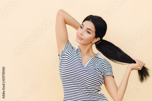 Fotografía Pensive dark haired female makes pony tail, has full lips and pleasant appearance, wears casual outfit, stands against studio background, focused into distance