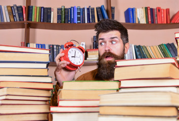 Leinwandbilder - Teacher or student with beard studying in library. Man, scientist peeking out of piles of books with alarm clock. Man on surprised face looking at clock, bookshelves on background. Deadline concept.