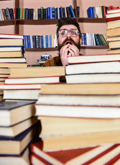 Leinwandbilder - Man on nervous face between piles of books in library, bookshelves on background. Teacher or student with beard wears eyeglasses, sits at table with books, defocused. Scientific discovery concept.