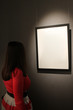 One blank white canvas frame to hang on the wall with the light shining down. The dark silhouette of a girl in a red dress looking at a picture or photo.