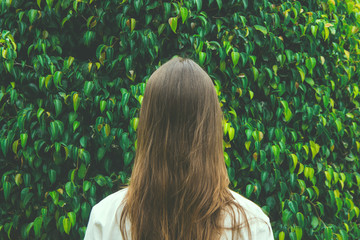 Wall Mural - Young Caucasian Woman Girl with Long Chestnut Hair Standing with Back to Viewer on Green Forest Tree Foliage Background. Contemplation Tranquility Mindful Leaving Wanderlust. Hipster Toned. Poster