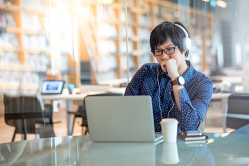 Wall Mural - Young Asian man listening to music working with laptop computer in public library. high school or university college student, educational concepts