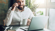© foxyburrow - Young smiling bearded businessman is sitting at table in front of laptop, drinking coffee, talking on cell phone. Telephone conversations, remote work, online training, education, marketing, working.