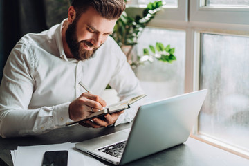 Young smiling bearded businessman in white shirt sitting at desk in front of laptop, making notes in notebook. Freelancer works remotely. Student learning online. Online traning, marketing, education.