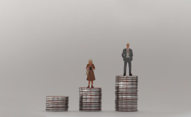 A miniature man and a miniature woman standing on a stack of coins of different heights. The concept of a gender pay gap.