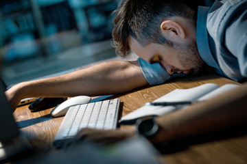 Young man working late in office and getting nap lying on hand at computer desk. 