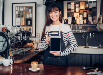 Poster - Asian woman barista wear jean apron holding blank screen tablet to customer at bar counter with smile emotion,Cafe restaurant service concept,Owner small business.mock up device for display text.