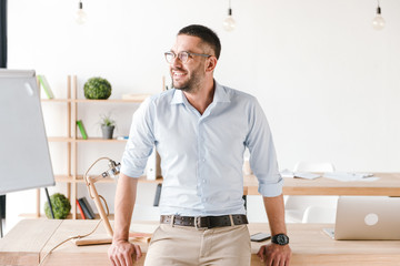 Happy portrait of businesslike man wearing white shirt sitting on table in office, and looking aside with smiling