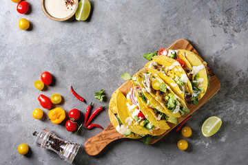 Variety of vegetarian corn tacos with vegetables, green salad, chili pepper served on olive wood board with tomato, cream sauces with ingredients above over grey texture background. Top view, space.