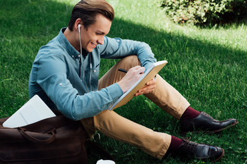 high angle view of smiling young man in earphones taking notes in notepad