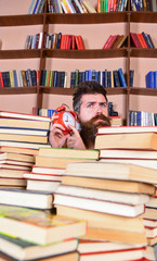 Leinwandbilder - Teacher or student with beard studying in library. Man, scientist peeking out of piles of books with alarm clock. Man on thoughtful face listening clock, bookshelves on background. Time stop concept.