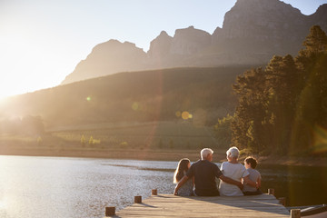 Grandchildren With Grandparents Sitting On Wooden Jetty By Lake