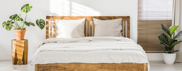 close-up of double wooden bed with bedding, pillows and blanket against white wall in a bright sunny