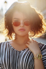 Outdoor close up portrait of young beautiful stylish curly girl wearing oval red color sunglasses, golden wrist watch, striped dress. Model posing in street. Sunny day light. Summer fashion concept